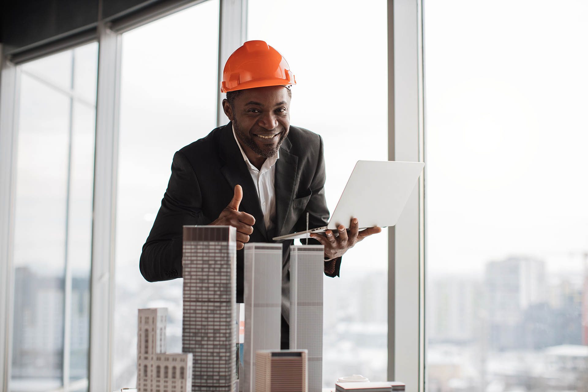 Happy african man in orange construction hard hat and suit showing thumb up and smiling on camera.
